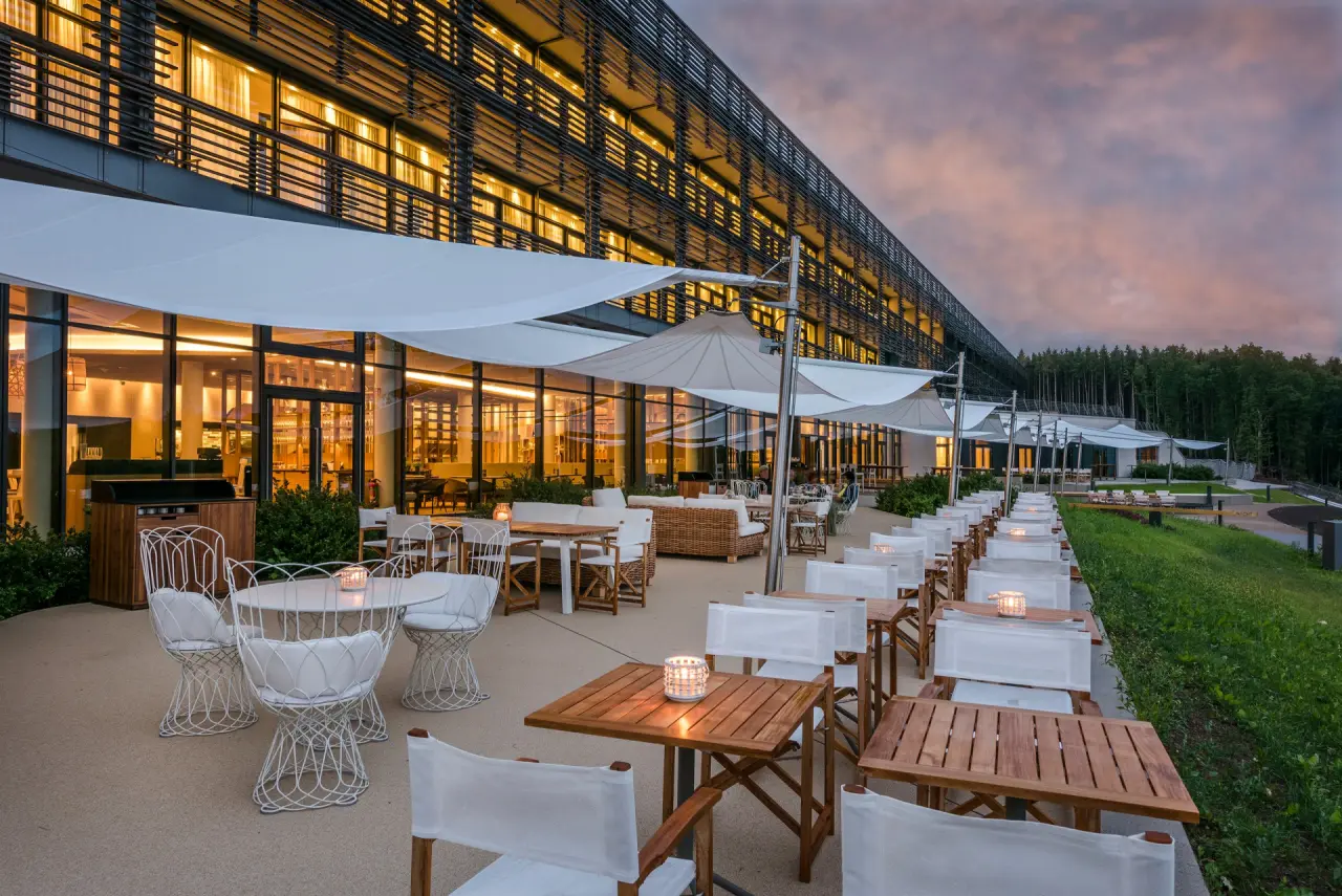 Abendliche Ansicht der Seezeitlodge mit beleuchteter Restaurantterrasse, modernen Möbeln und Waldkulisse bei Dämmerung.
