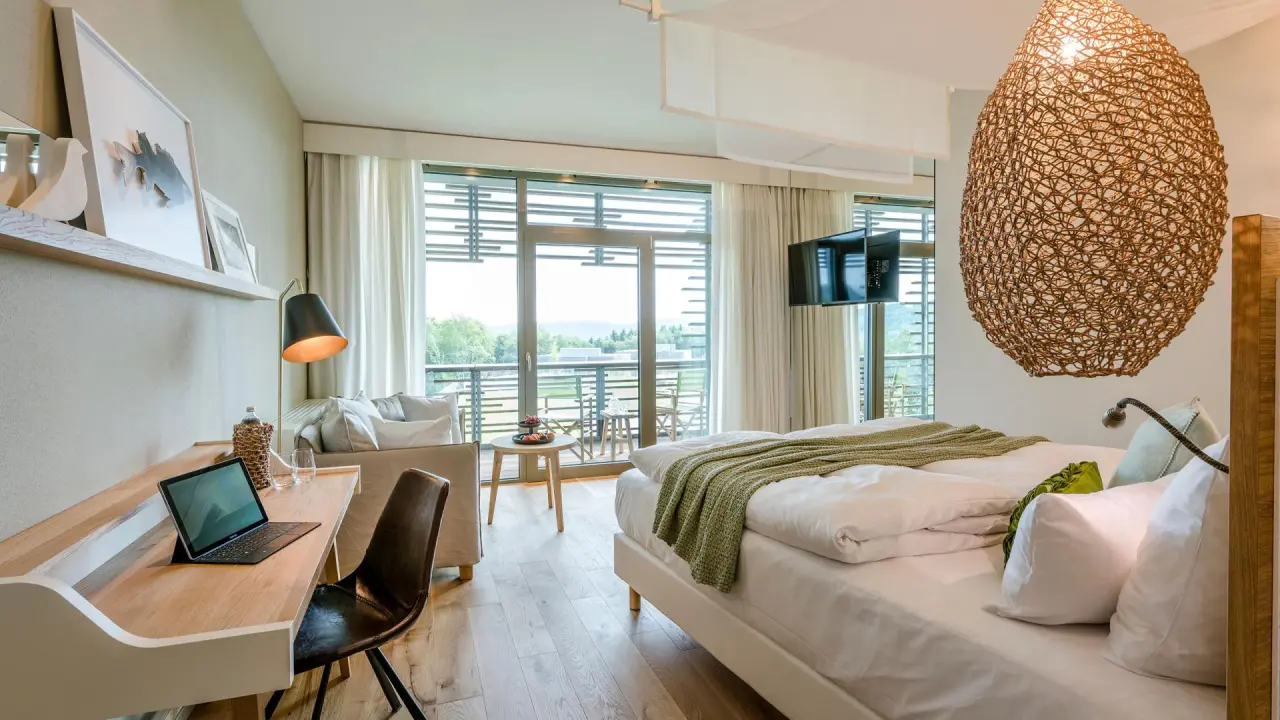 Modernes Hotelzimmer der Seezeitlodge mit Balkon, Schreibtisch, Doppelbett und Ausblick ins Grüne.