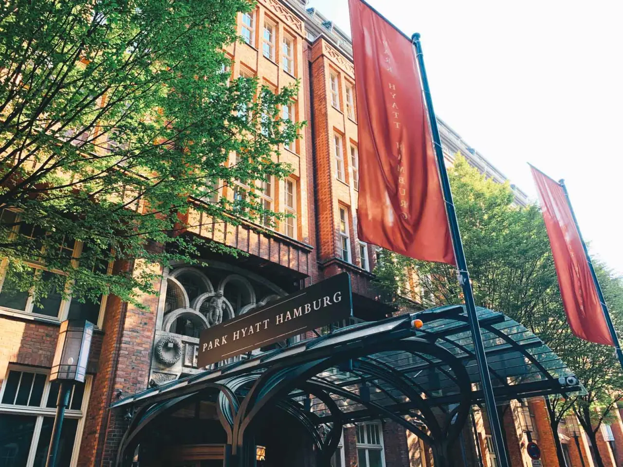 Historische Backsteinfassade und Eingangsbereich des Park Hyatt Hamburg Hotels mit roten Bannern und grünen Bäumen.