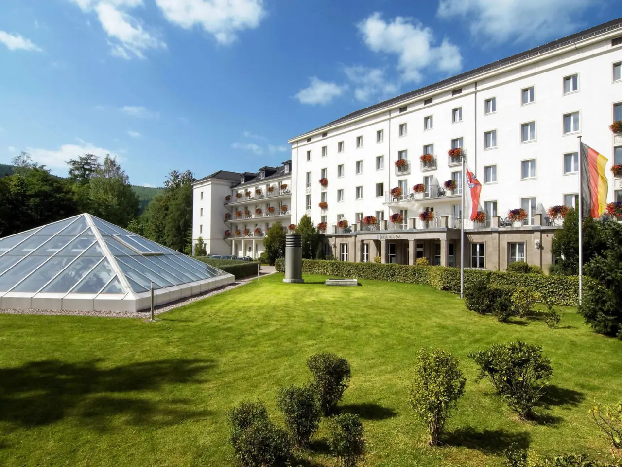 Aussenansicht des H+ Hotel & SPA Friedrichroda mit weisser Fassade, Balkonen, gruenem Garten und Glaspyramide unter blauem Himmel.