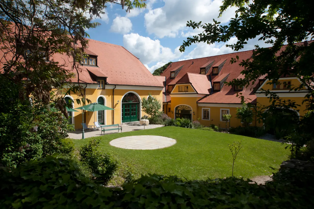 Sonniger Innenhof mit gelben Gebäuden und rotem Dach des Hotel Althof Retz, umgeben von grünem Garten und Bäumen.
