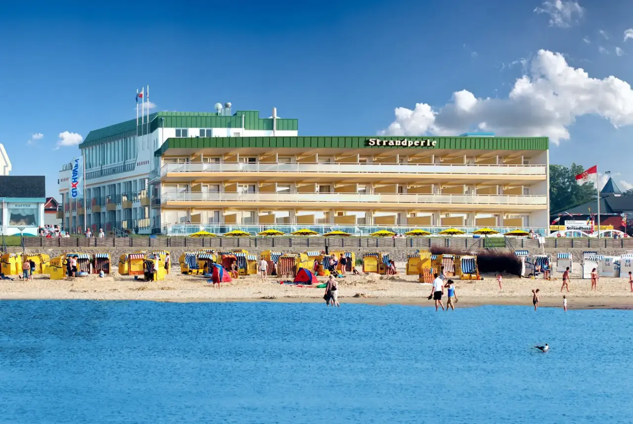 Hotel Strandperle direkt am Strand mit Meerblick, Strandkörben und Badegästen unter blauem Himmel.