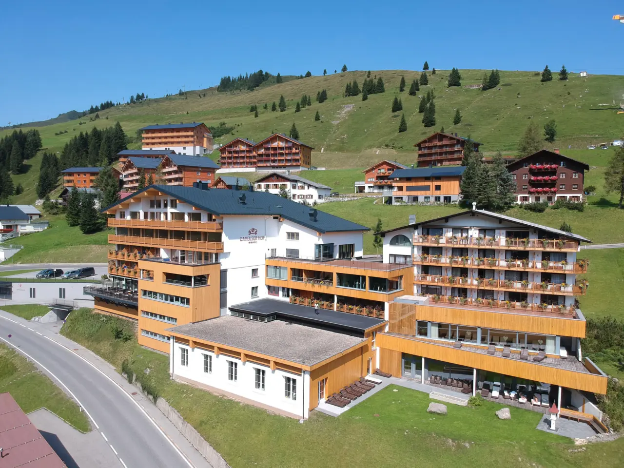 Luftaufnahme des Damülser Hof Hotels in den österreichischen Alpen. Mehrere Holzgebäude mit Balkonen vor grünen Berghängen.