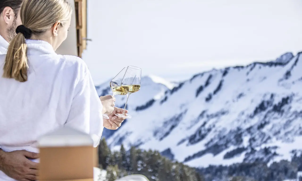 Paar im Bademantel genießt Wein auf dem Balkon mit Blick auf die verschneiten Berge im Damülser Hof.