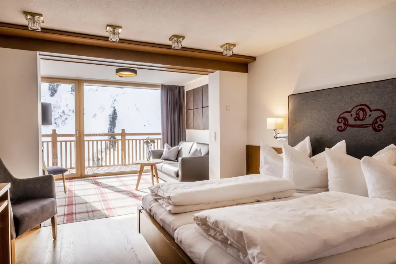 Gemütliches Hotelzimmer im Damülser Hof mit Doppelbett, Sitzbereich und Balkon mit Blick auf die verschneite Berglandschaft.