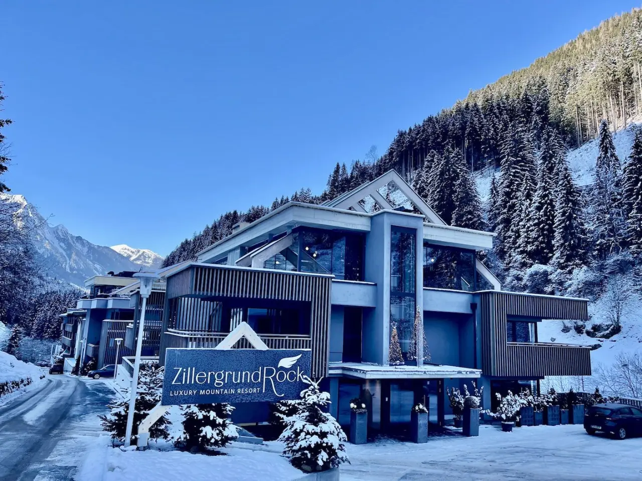 Modernes ZillergrundRock Luxury Mountain Resort im Winter mit verschneiten Bergen und Wald unter blauem Himmel.