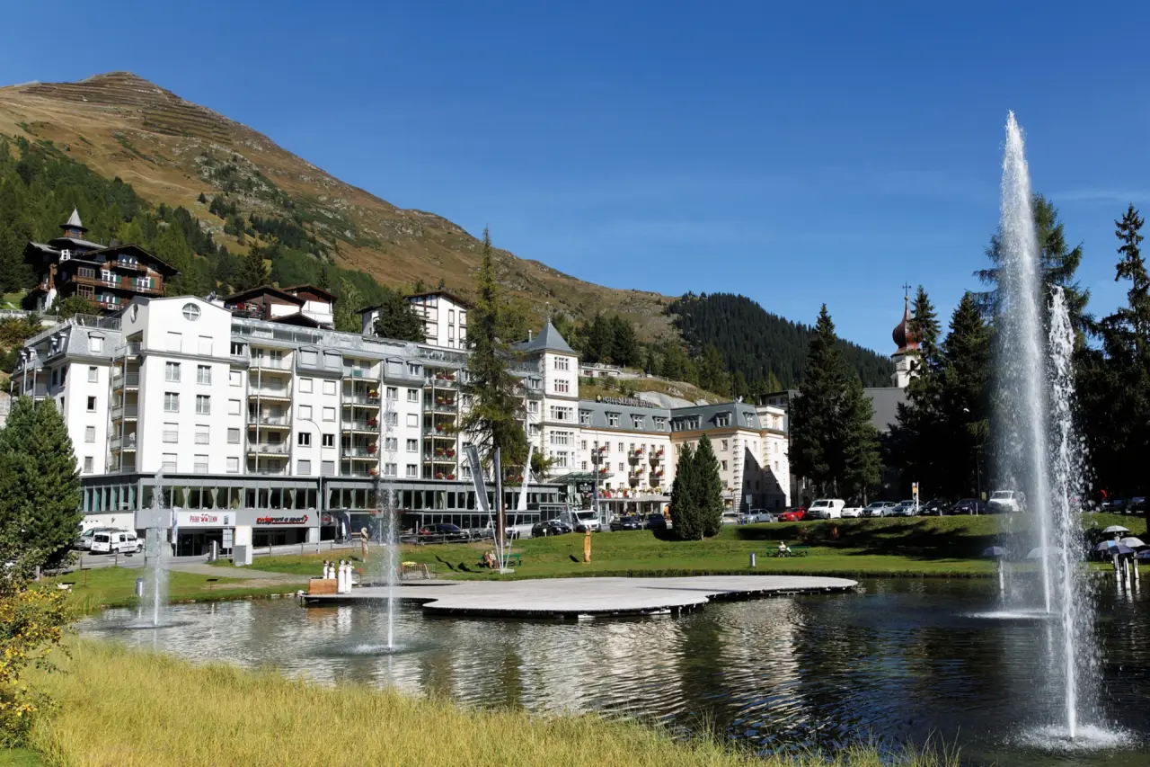Aussenansicht des Precise Tale Seehof Davos mit Bergpanorama und Teich mit Springbrunnen an einem sonnigen Tag.