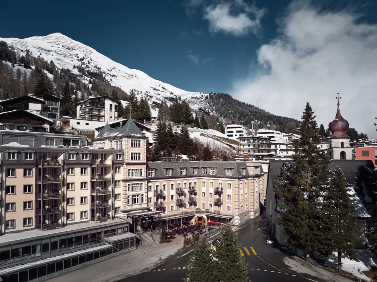 Majestätische Aussenansicht des Precise Tale Seehof Davos im Winter, umgeben von schneebedeckten Schweizer Alpen unter blauem Himmel.