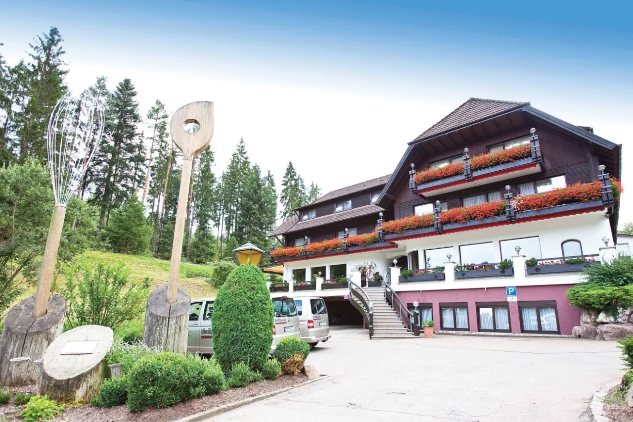 Aussenansicht Hotel Waldsägmühle mit traditioneller Architektur, roten Blumen und grossen Kochutensilien im Vorgarten.