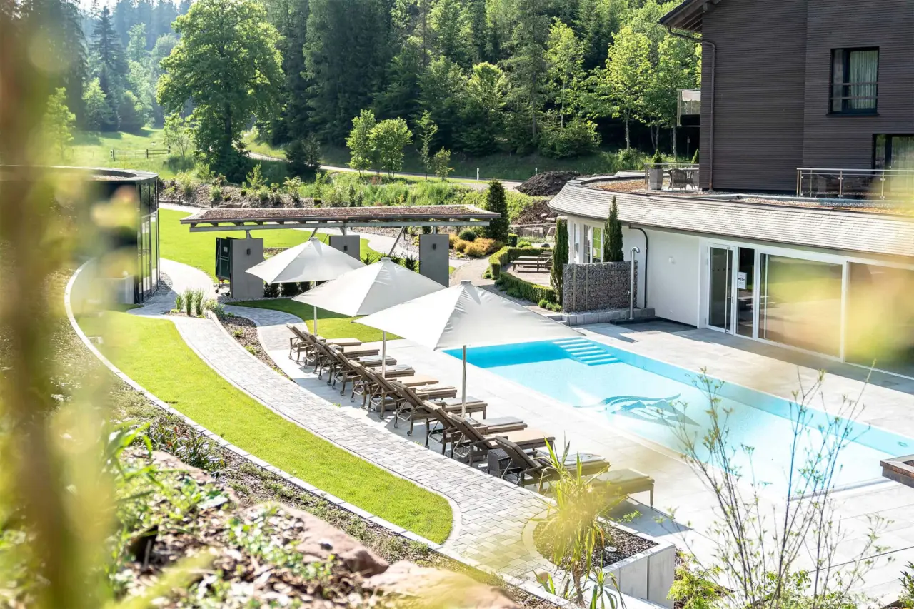 Aussenpool mit Liegen und Sonnenschirmen im grünen Garten des Hotel Waldsägmühle, umgeben von Wald.