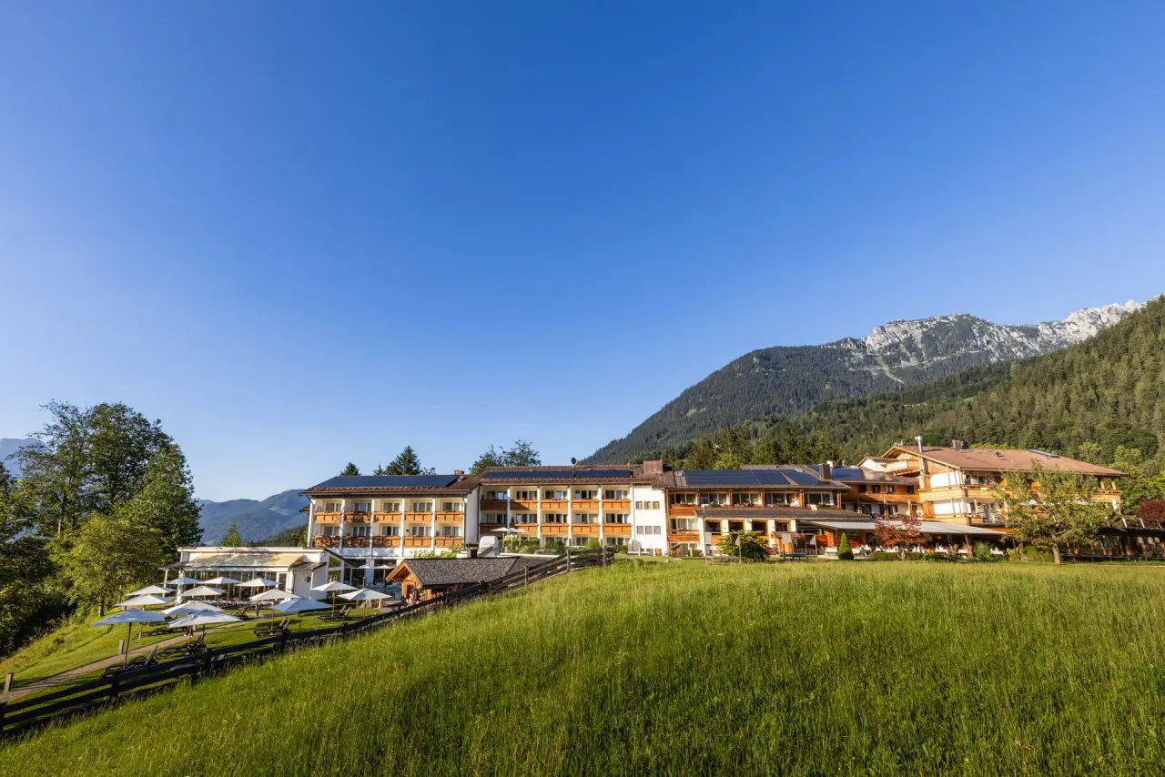 Alm- & Wellnesshotel Alpenhof in den Bergen, umgeben von grünen Wiesen und Wäldern unter blauem Himmel.