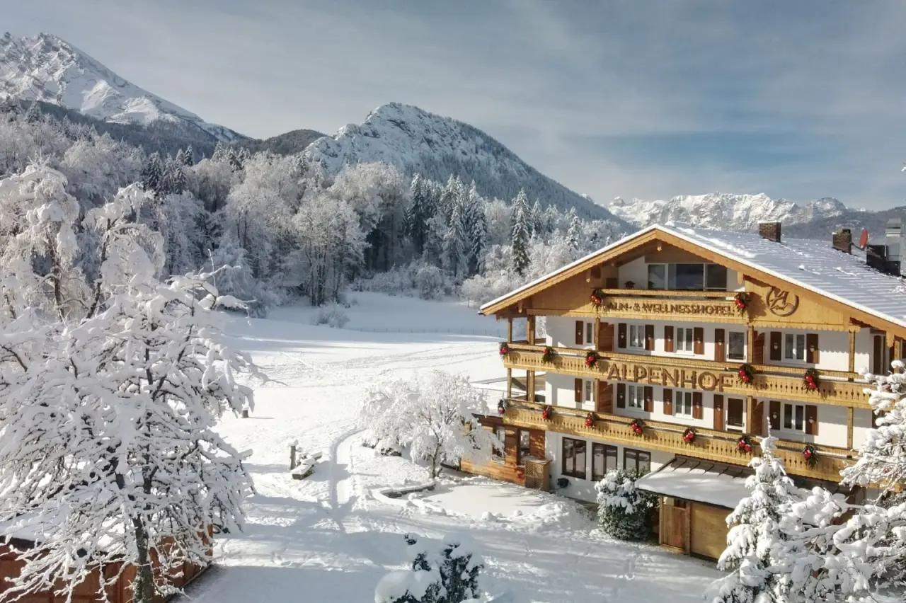 Alm- & Wellnesshotel Alpenhof im Winter, umgeben von verschneiten Bergen und Wäldern unter blauem Himmel.