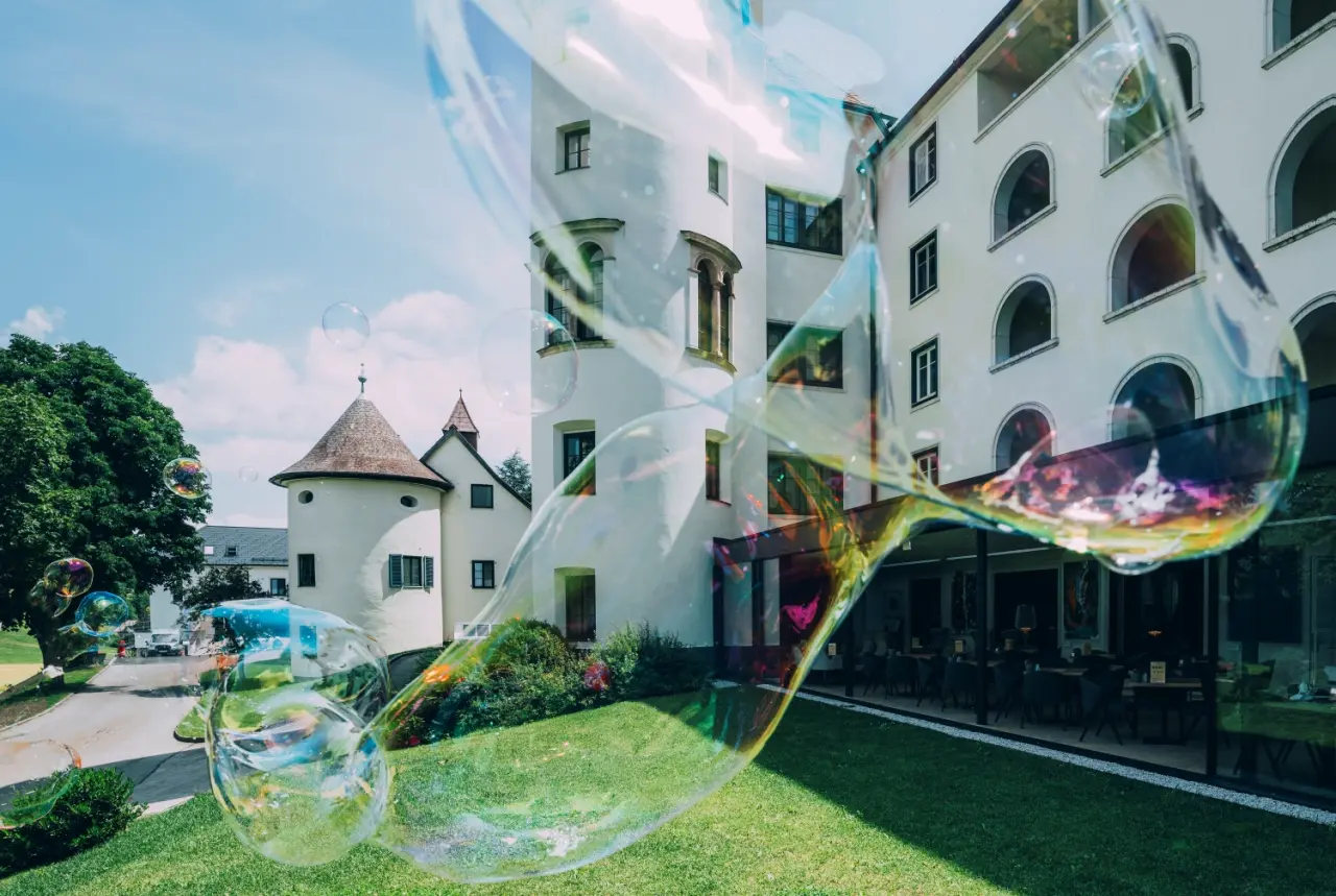 Historisches IMLAUER Hotel Schloss Pichlarn mit weisser Fassade, Türmchen und grünem Garten unter blauem Himmel, mit Seifenblasen.