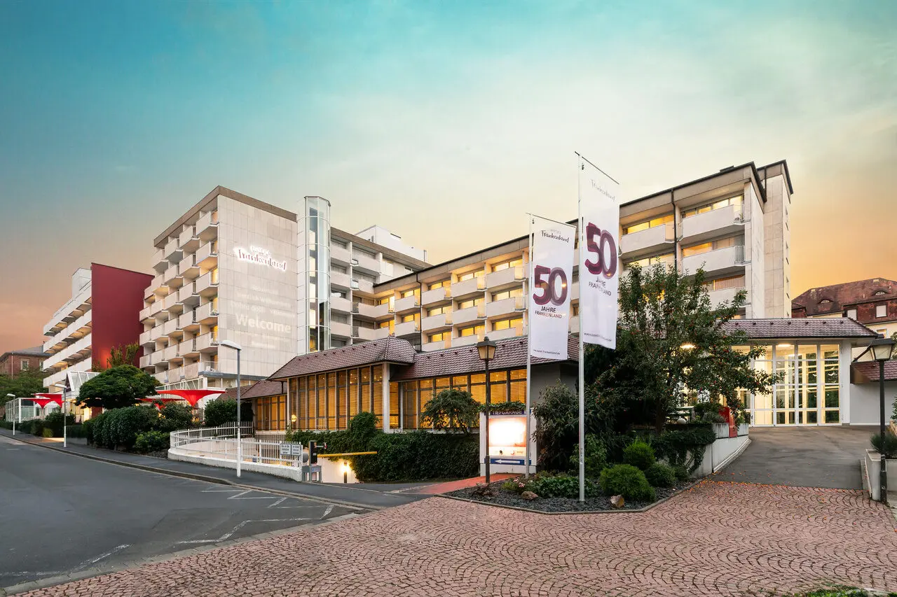 Aussenansicht des Hotel Frankenland bei Sonnenuntergang mit beleuchteten Fenstern und '50 Jahre' Flaggen.