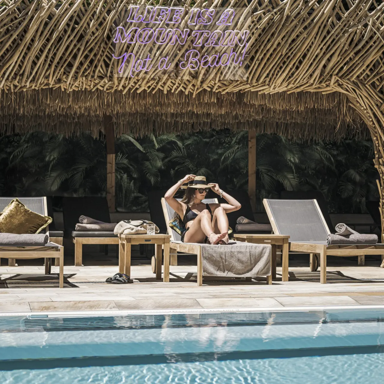 Gast entspannt am Aussenpool des Alpine Hideaway Zillertalerhof unter einem Strohdach mit Neon-Schriftzug.