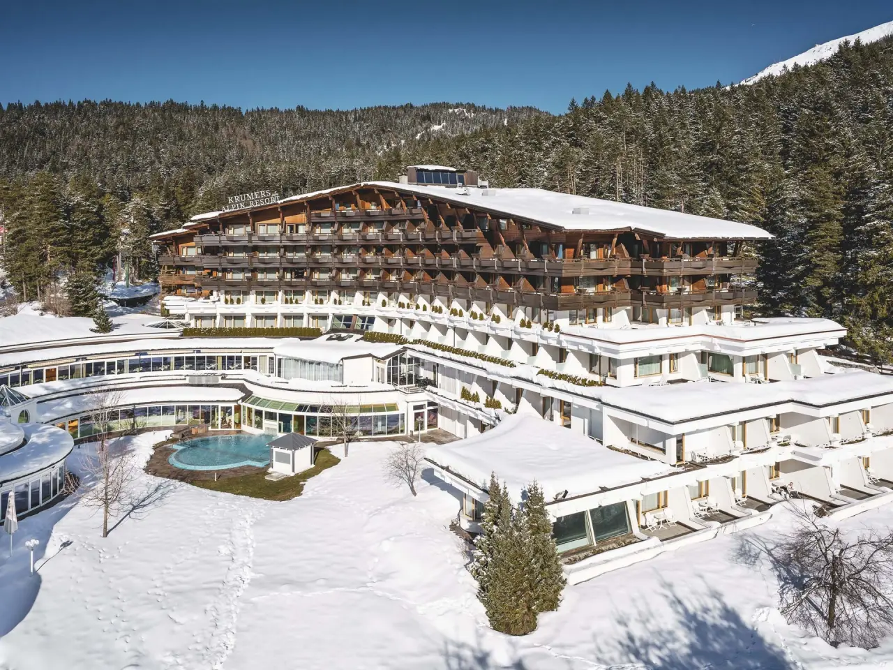 Winterliche Aussenansicht des Krumers Alpin Resort mit verschneitem Hotelgebäude und beheiztem Aussenpool vor Bergkulisse.