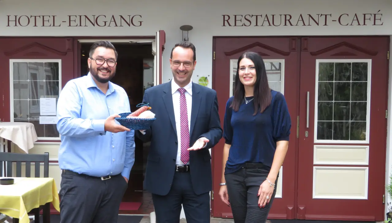 Drei lächelnde Mitarbeiter des Landhotel Kern vor dem Hoteleingang und Restaurant-Café, einer hält einen Korb mit Wurst.