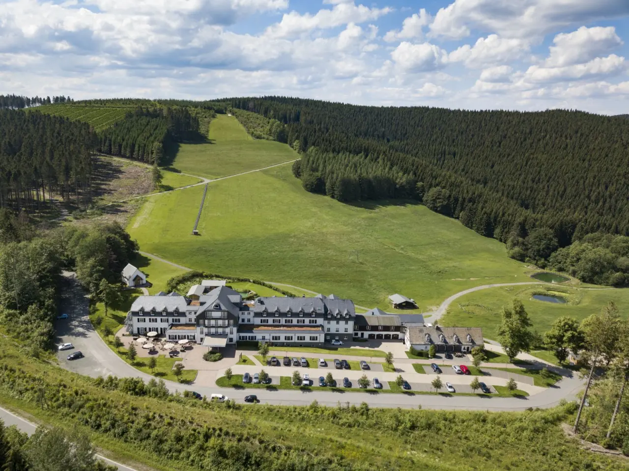 Luftaufnahme des Hotel Rimberg inmitten grüner Wiesen und Wälder im Sauerland, mit Parkplätzen und Sonnenterrasse.