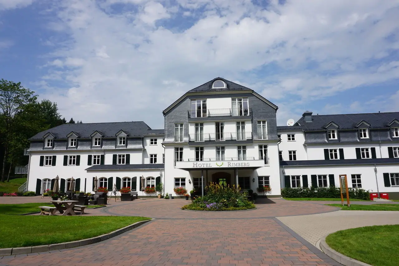 Elegante Aussenansicht des Hotel Rimberg mit weisser Fassade, Schieferdach und blühendem Vorgarten an einem sonnigen Tag.