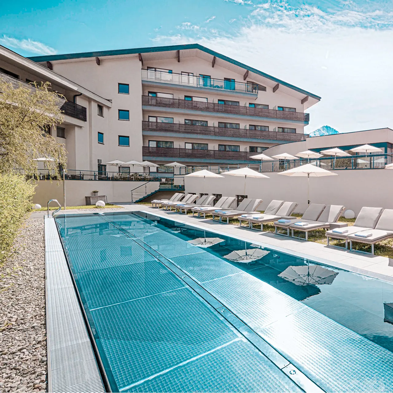 Grosszügiger Aussenpool mit Liegen und Hotelgebäude des HAIDVOGL MAVIDA Zell am See unter blauem Himmel.