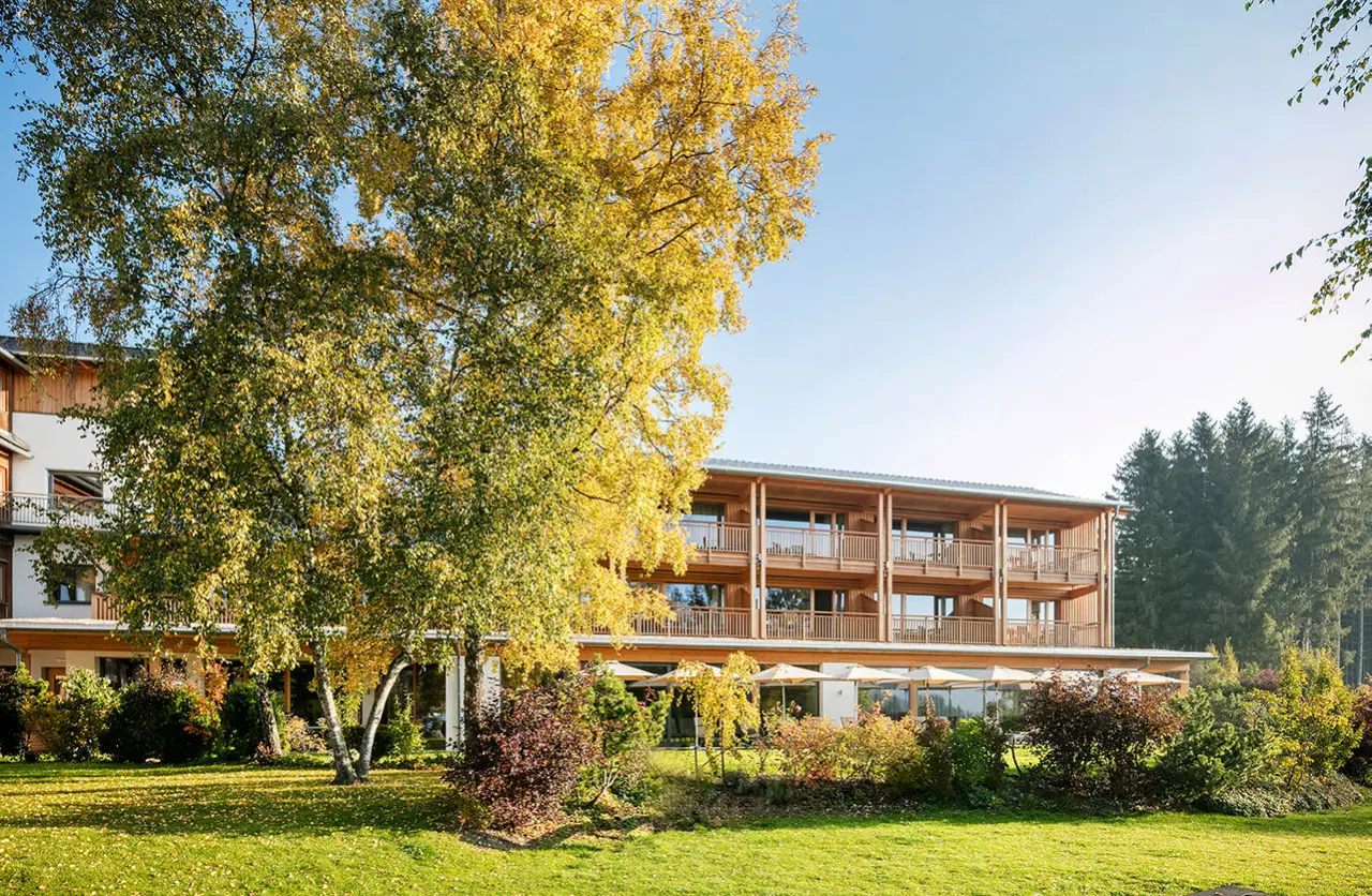 Aussenansicht des Naturhotel Bauernhofer im Herbst mit goldgelben Birken und grüner Wiese unter blauem Himmel.