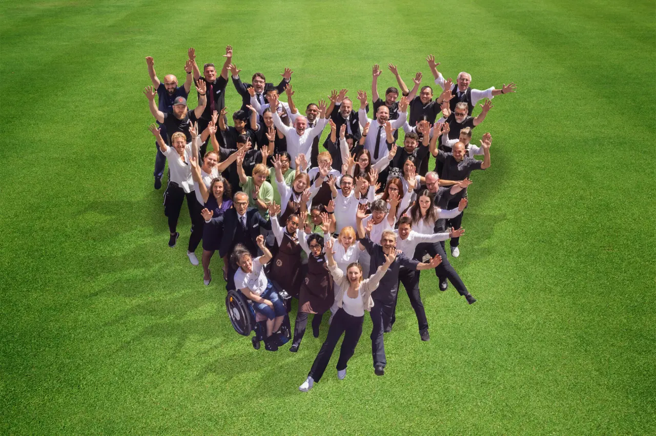 Das freundliche Team des Schwarzwald Panorama Hotels in Herzform auf einer grünen Wiese, herzlich winkend.