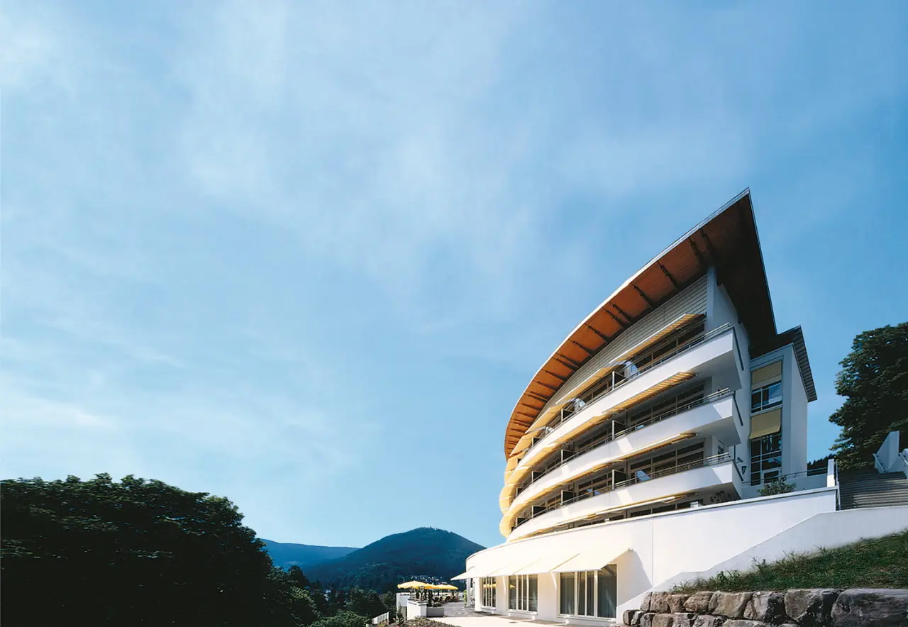 Modernes Hotel Schwarzwald Panorama mit geschwungener Architektur und Balkonen, eingebettet in die grüne Landschaft des Schwarzwaldes.
