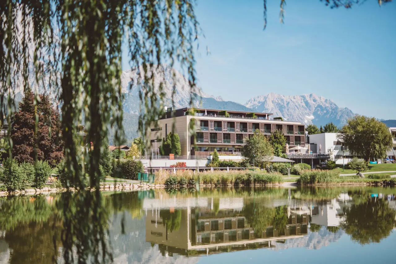 Modernes Hotelgebäude des Ritzenhof****S am See mit spiegelnder Wasseroberfläche und majestätischem Bergpanorama im Hintergrund.