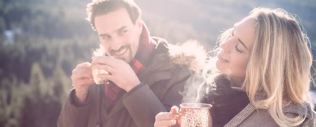 Lachendes Paar genießt dampfende Heißgetränke im winterlichen Freien des Naturhotel Edelweiss.