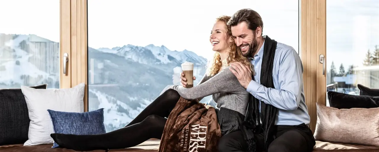 Glückliches Paar entspannt auf einer Fensterbank mit Bergblick im Naturhotel Edelweiss, genießt Kaffee und die Alpenkulisse.