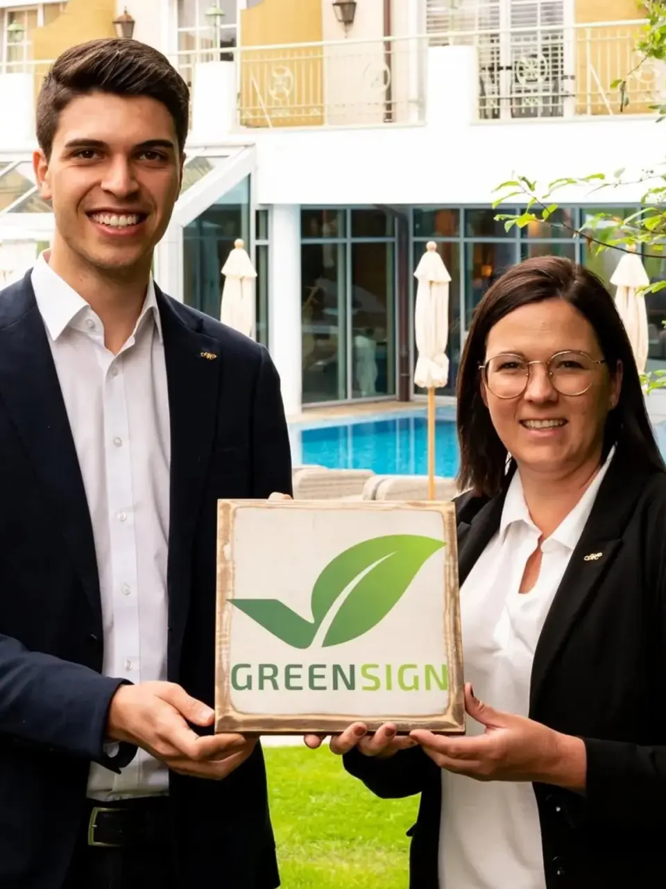 Mitarbeiter des Meiser Vital Hotels präsentieren stolz das GREENSIGN Nachhaltigkeitssiegel vor der Poolanlage.
