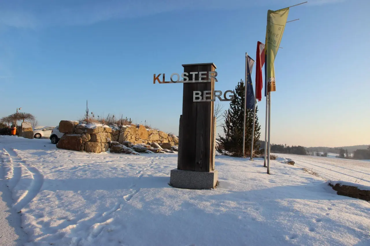 Eingangsschild des Gesundheitshotels Klosterberg im Winter mit verschneiter Landschaft und blauem Himmel.