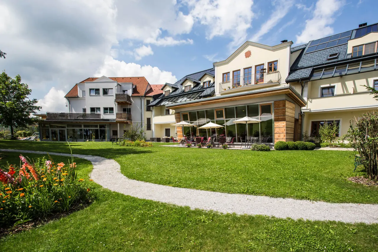 Aussenansicht des Gesundheitshotels Klosterberg mit gepflegtem Garten, Terrasse und blühenden Blumen unter blauem Himmel.