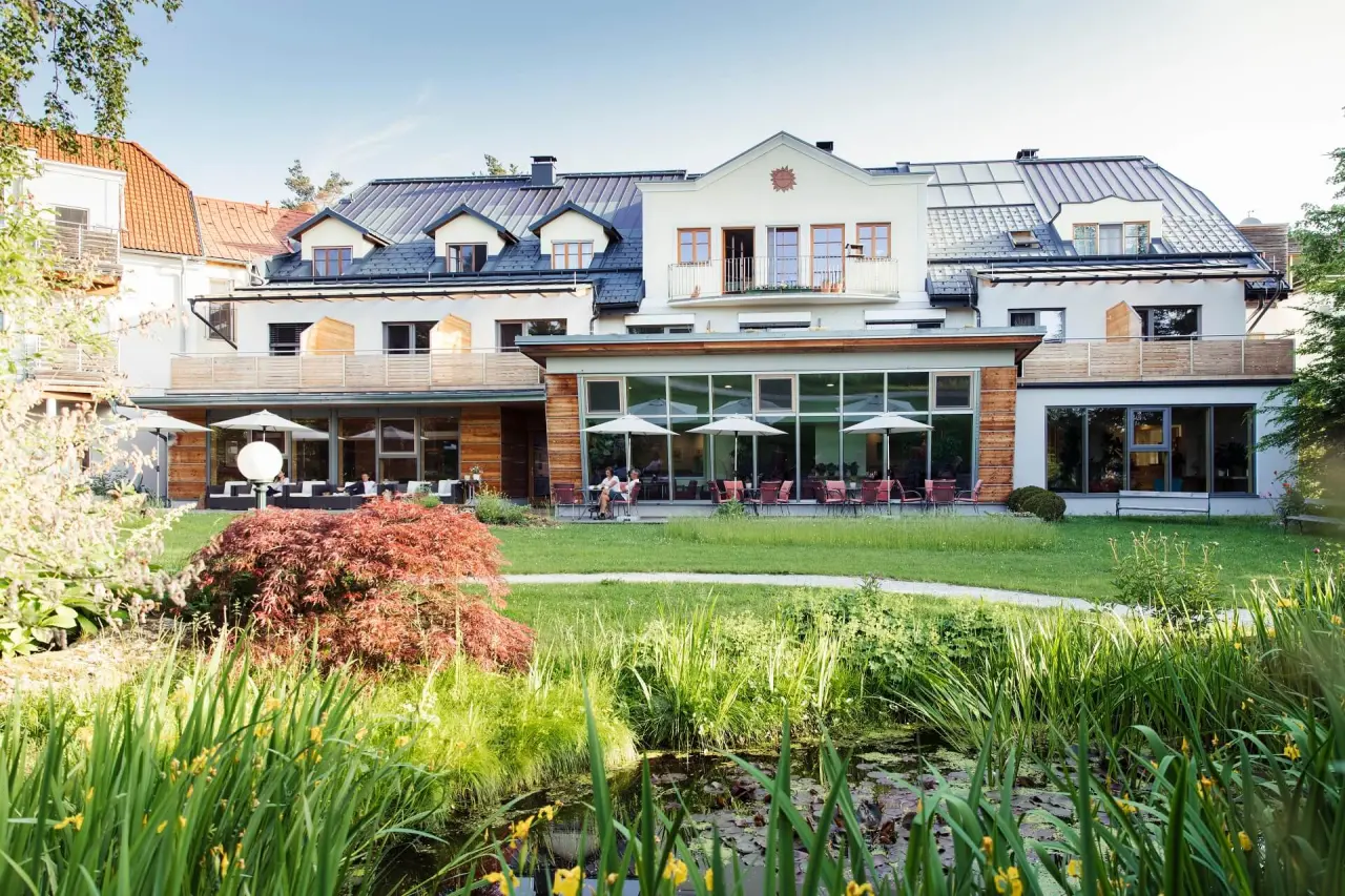 Aussenansicht des Gesundheitshotels Klosterberg mit gepflegtem Garten, Terrasse und Teich an einem sonnigen Tag.