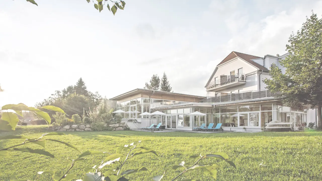 Aussenansicht des Gesundheitshotel Klosterberg mit moderner Glasfassade, Liegestühlen und weitläufiger Grünfläche im Sonnenlicht.