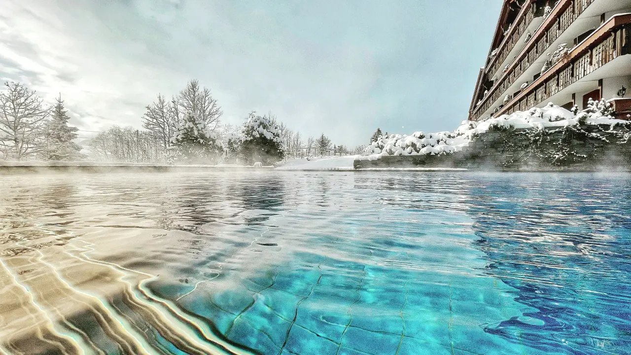 Beheizter Aussenpool mit Dampf im Schnee und Hotelgebäude des ERMITAGE Wellness- & Spa-Hotel.