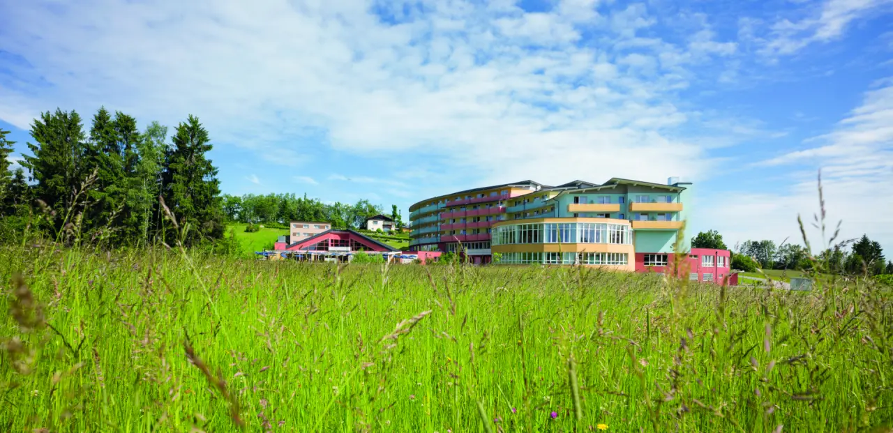 Aussenansicht des Vivea 4* Hotel Bad Traunstein in Bad Traunstein, umgeben von grünen Wiesen und Wäldern unter blauem Himmel.