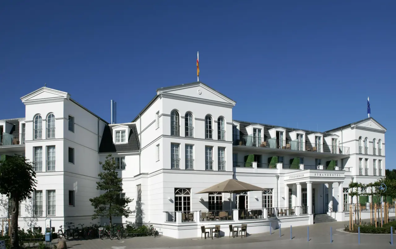 Elegantes weißes Strandhotel Zingst mit klassischer Architektur und Außenterrasse unter blauem Himmel.
