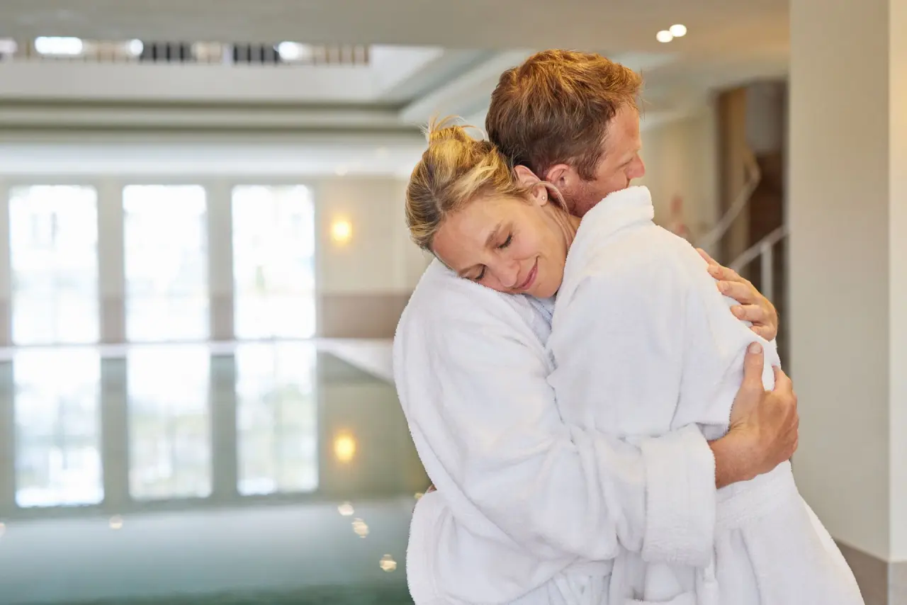 Entspanntes Paar in Bademänteln umarmt sich im Wellnessbereich des Strandhotel Zingst, mit Pool im Hintergrund.
