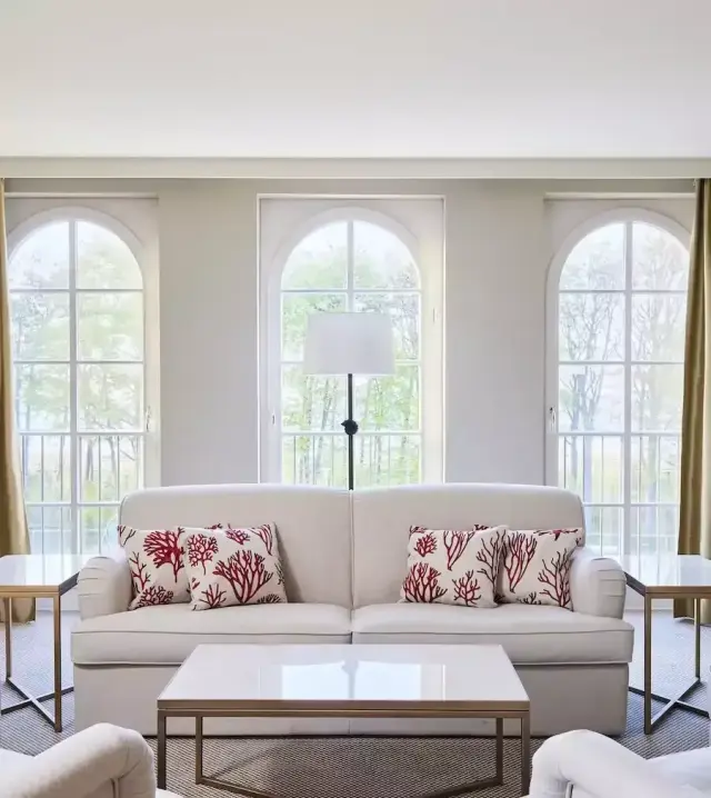 Helles Wohnzimmer im Strandhotel Zingst mit weissem Sofa, Korallenkissen und grossen Bogenfenstern mit Blick ins Grüne.