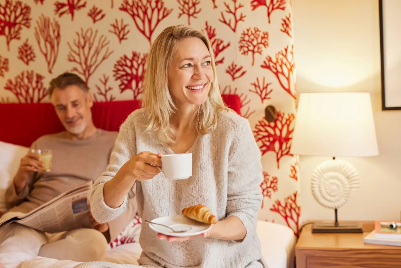 Glückliches Paar beim Frühstück im Bett in einem komfortablen Zimmer des Strandhotel Zingst mit maritimem Dekor.
