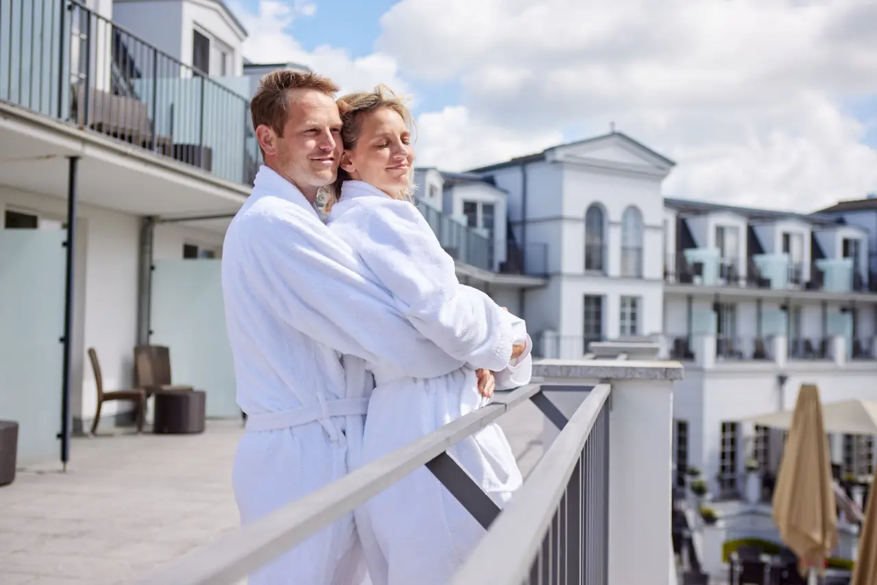 Entspanntes Paar in Bademänteln auf dem Balkon des Strandhotel Zingst mit Blick auf die Hotelarchitektur.