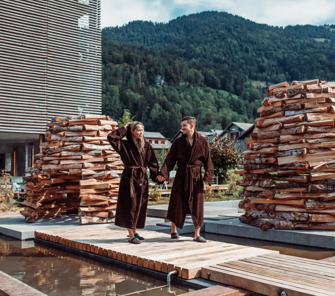 Paar in Bademänteln auf Holzsteg über Wasser mit Holzstapeln und Bergkulisse im Wellnesshotel GAMS zu zweit.