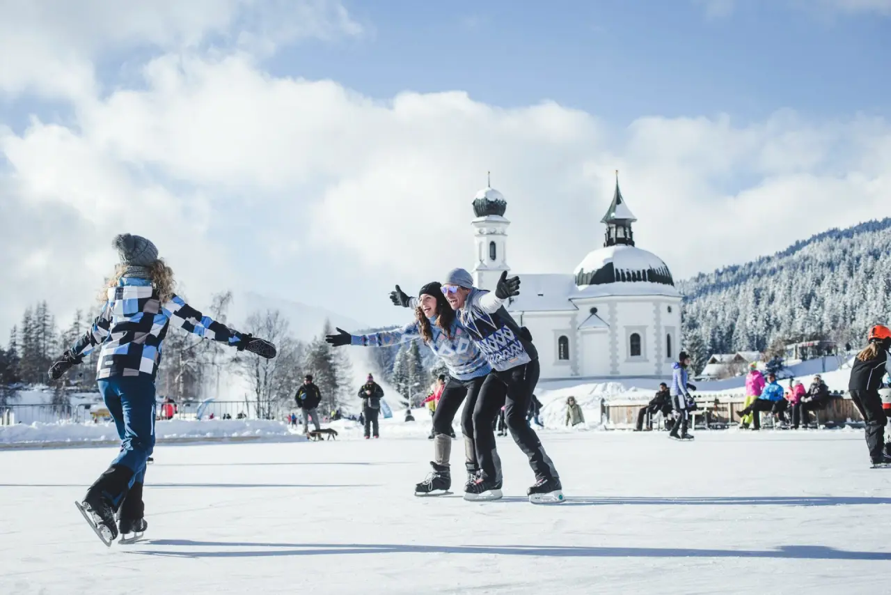 Fröhliche Eisläufer auf einer Natureisbahn mit verschneiter Kirche und Bergen im Hintergrund, nahe Hotel Eden.