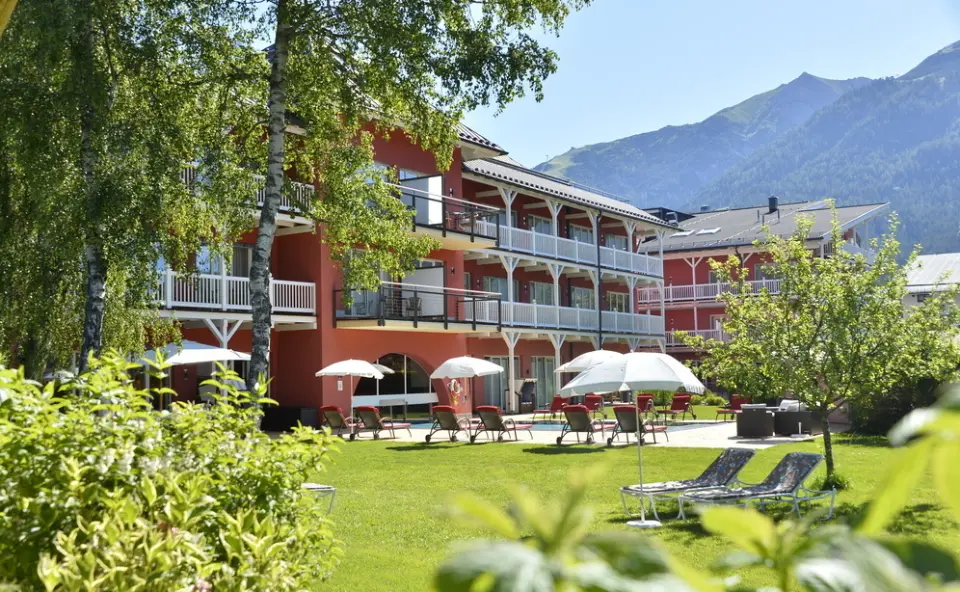 Aussenansicht des Hotel Eden mit roter Fassade, weissen Balkonen, sonnigem Garten, Liegestühlen und Bergen im Hintergrund.