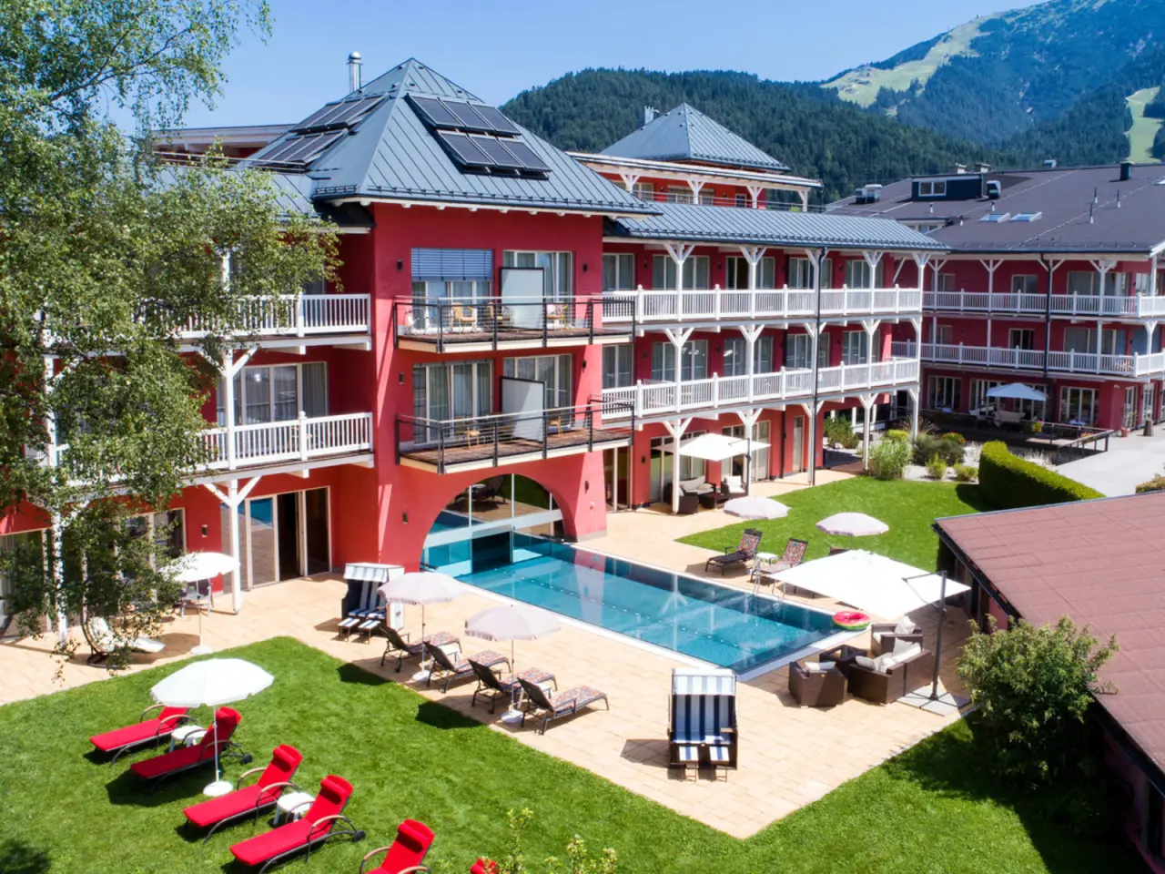 Aussenansicht des Hotel Eden mit roter Fassade, grossem Aussenpool und Liegewiese vor Bergkulisse.