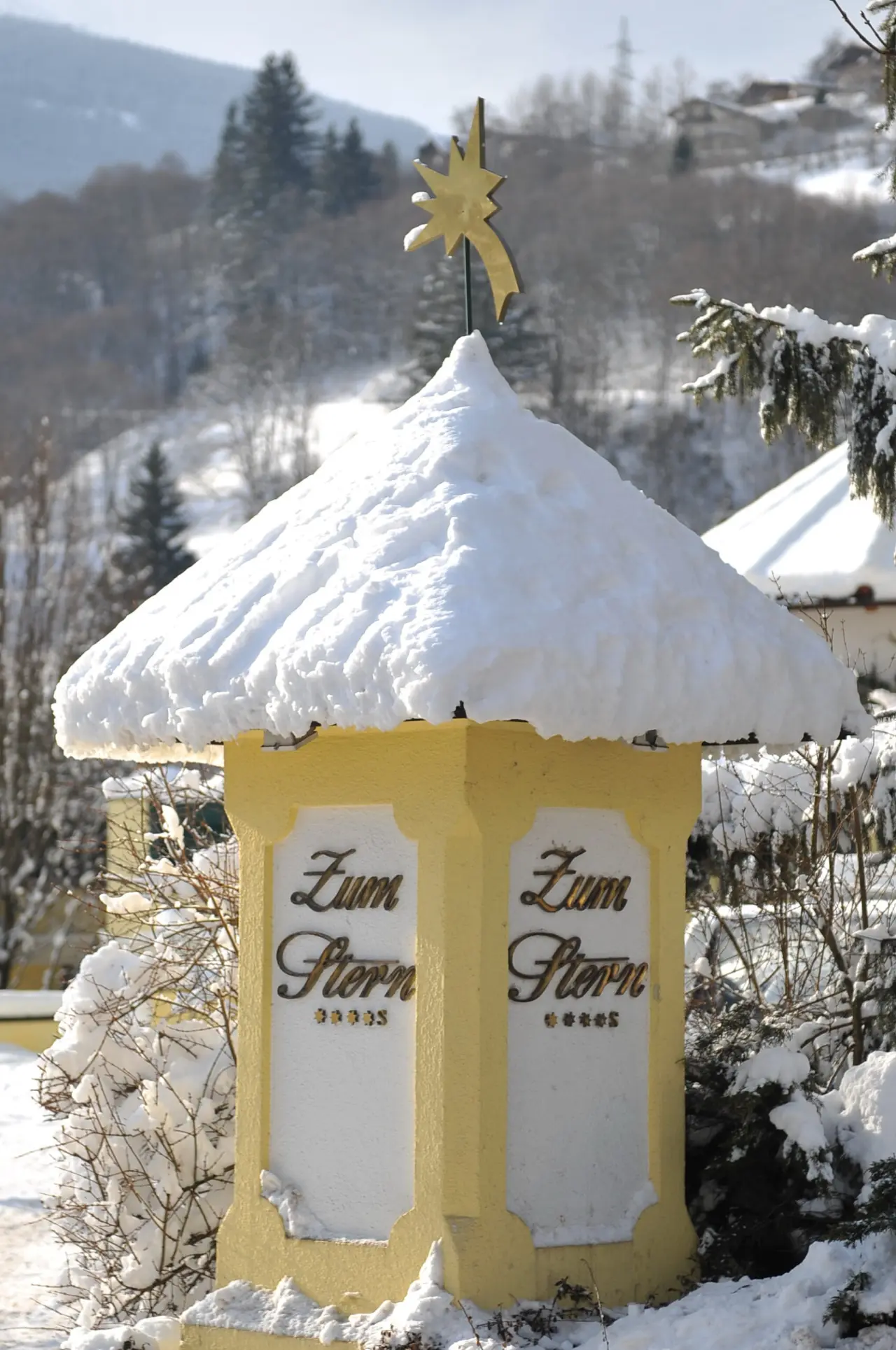 Schneebedeckter gelber Pfeiler mit 'Zum Stern' Schriftzug und goldenem Stern im winterlichen Garten des Sonnenhotel ZUM STERN.