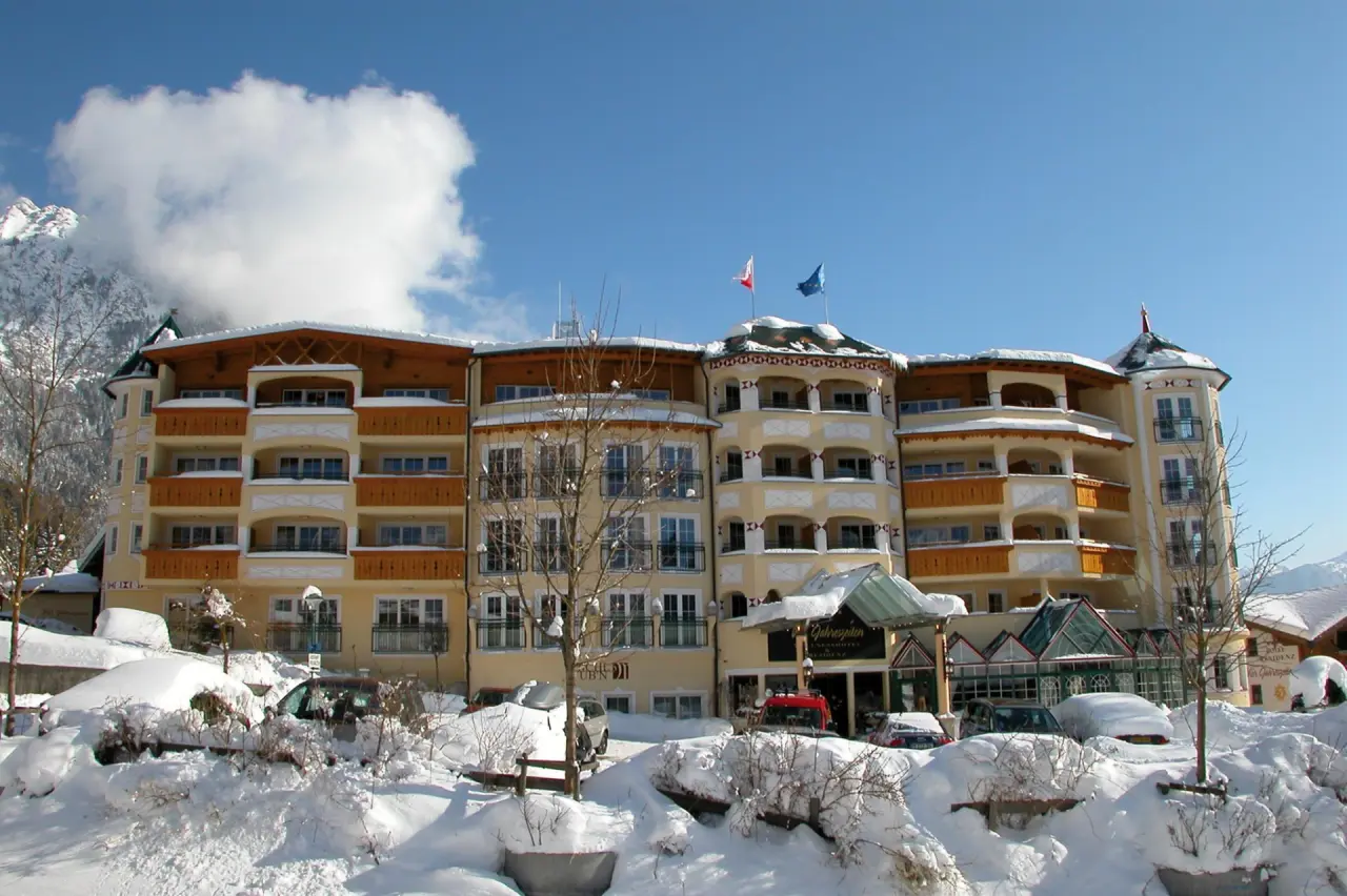 Winterliche Aussenansicht des Hotel Vier Jahreszeiten mit verschneiter Landschaft und Bergen im Hintergrund unter blauem Himmel.