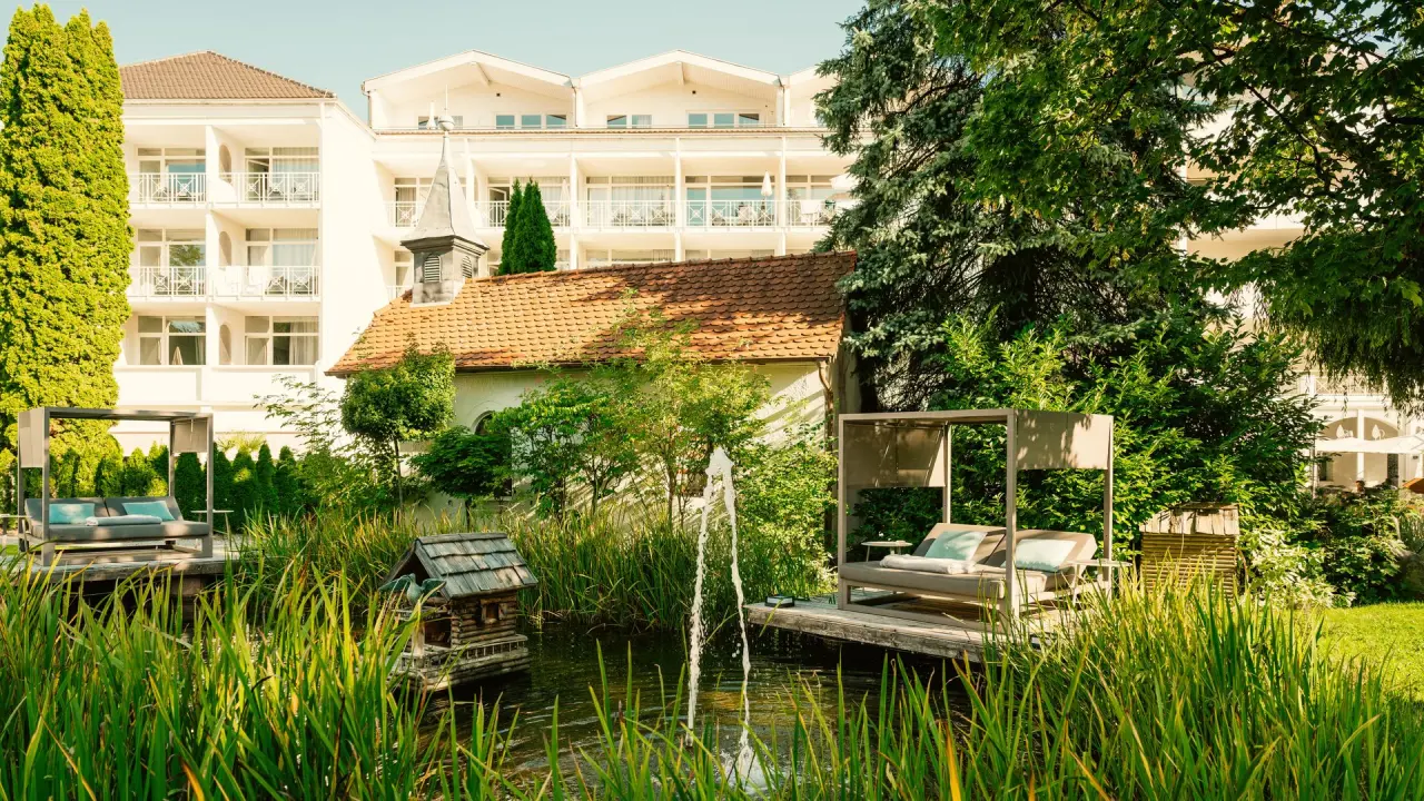 Entspannende Daybeds am Teich im grünen Garten des Parkhotels mit Hotelgebäude im Hintergrund.