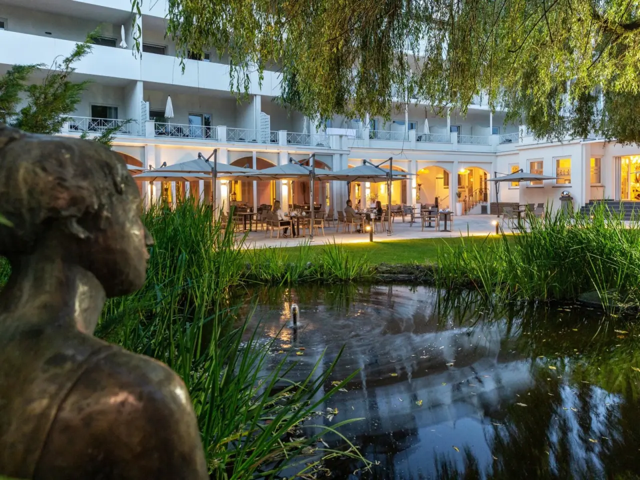 Abendliche Aussenansicht des Parkhotels mit beleuchteter Terrasse, Teich und Gartenbereich.